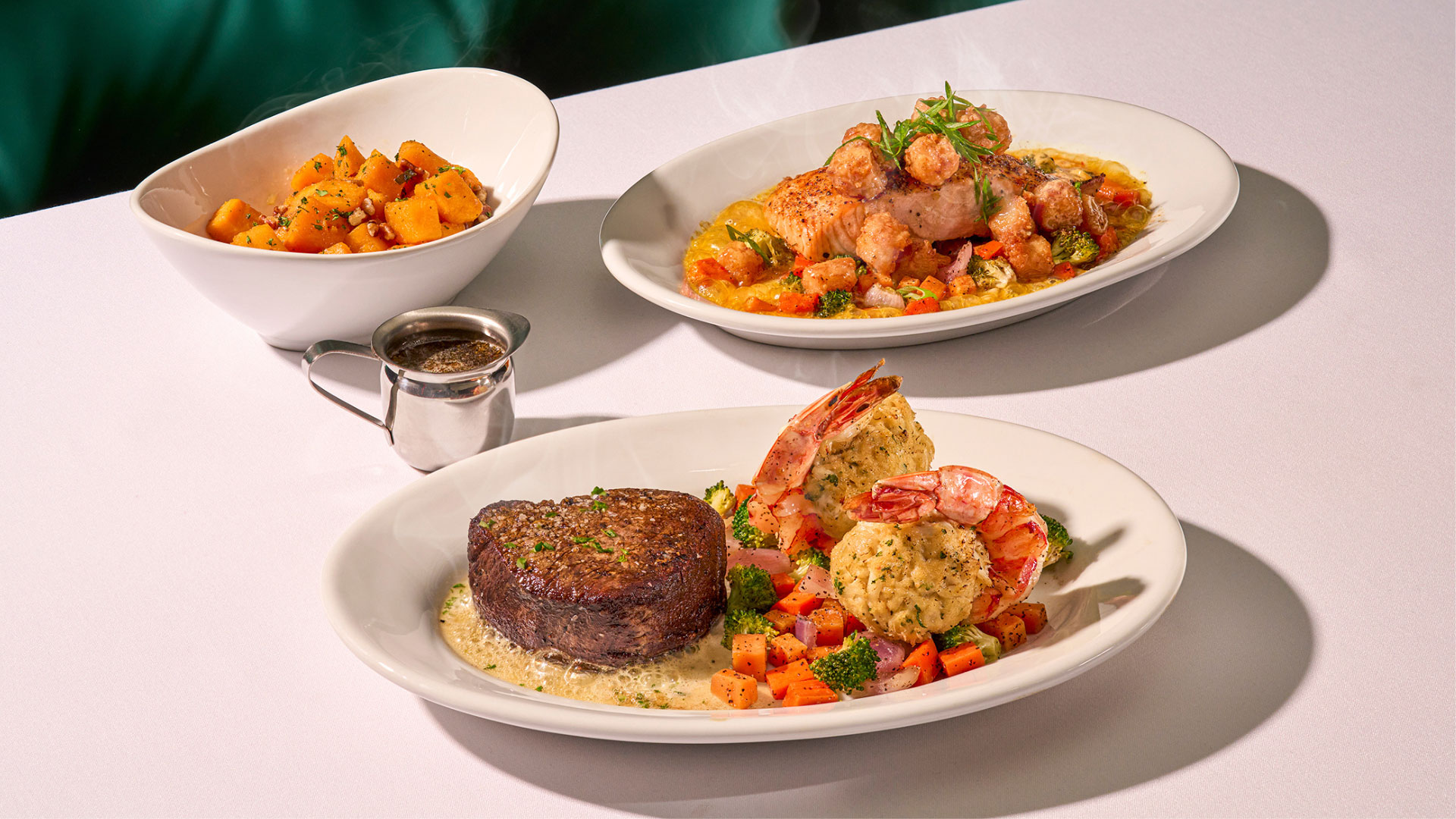 Three plates from Ruth's Chris' Chef Selected Items served on a table, including the center-cut filet and crab stuffed shrimp, the salmon with bay scallops, and maple-glazed butternut squash. 